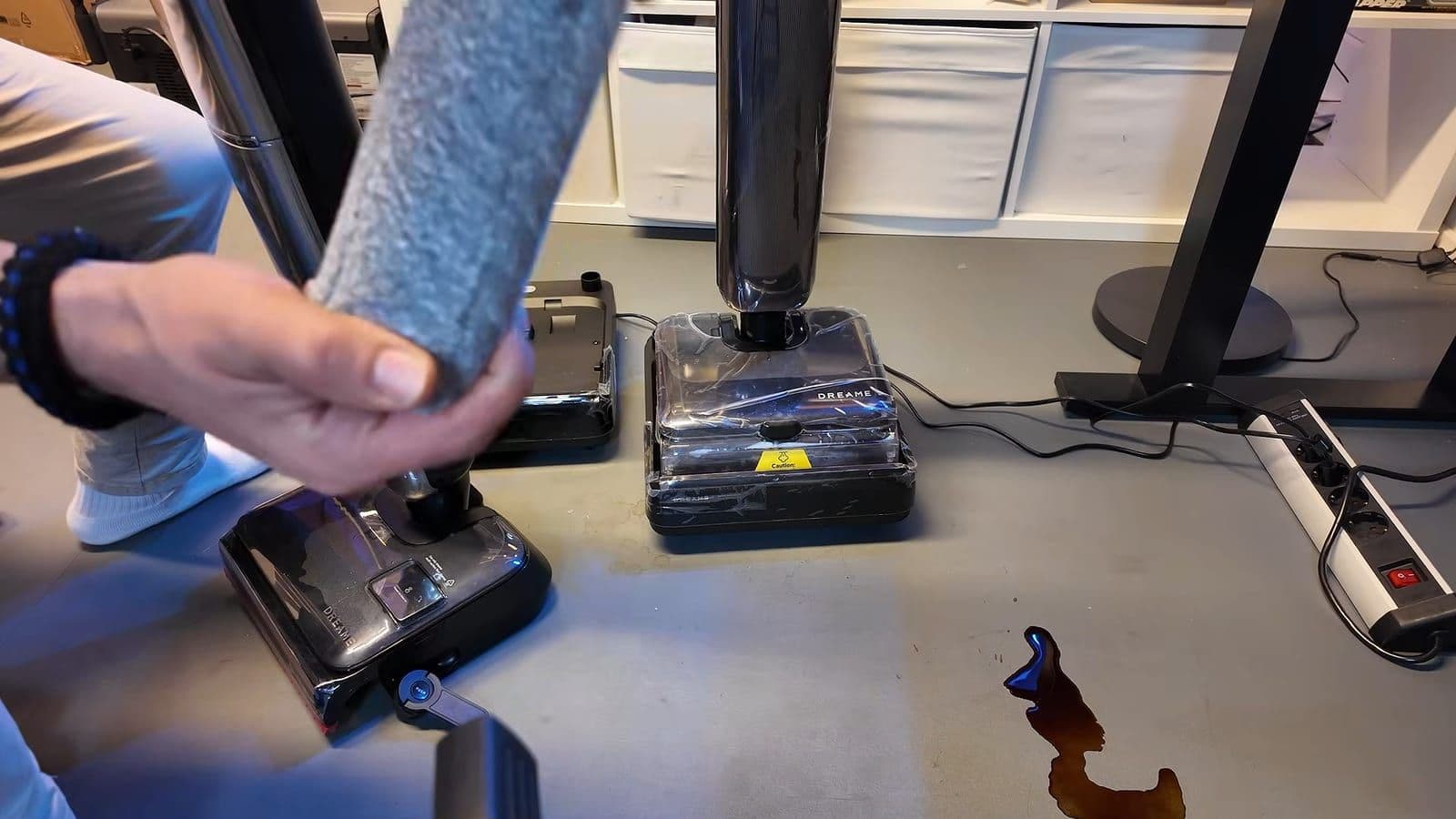 Een hand houdt een grijze microvezelroller omhoog boven de reinigingsstations van beide Dreame-stofzuigers
