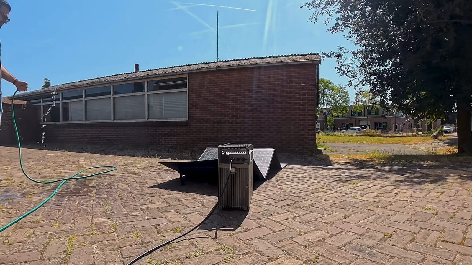 Een hand met een groene tuinslang spuit water over de draaiende batterij die buiten op de stenen staat