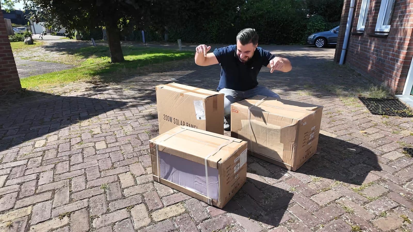Zino gehurkt op een bestrate oprit naast drie kartonnen dozen, waarvan één met de tekst 200W Solar Panel